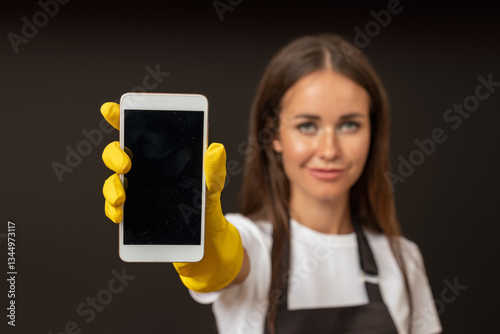 A female cleaner in a black apron and yellow rubber gloves, holding out a smartphone towards the camera, standing against a dark background.