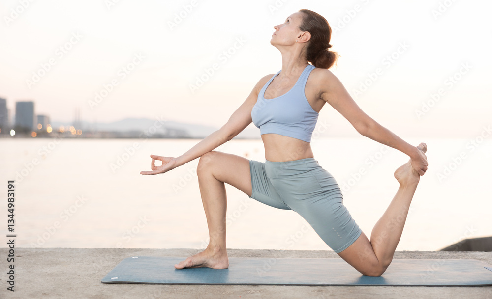 Fototapeta premium Young attractive smiling woman wearing sportswear practicing yoga, sitting in One Legged King Pigeon exercise, Eka Pada Rajakapotasana pose, working out on beach at sunset outdoors
