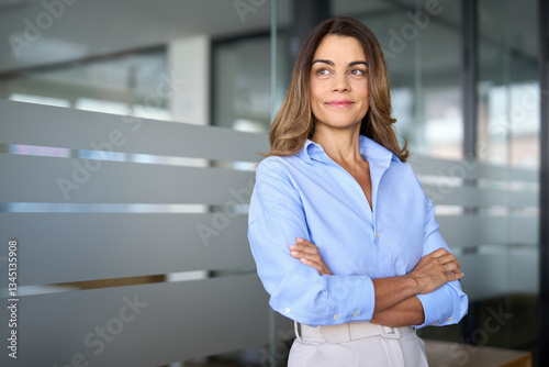 Smiling successful mature businesswoman of middle age standing in office arms crossed. Happy confident older professional business woman leader executive looking away thinking of future success.