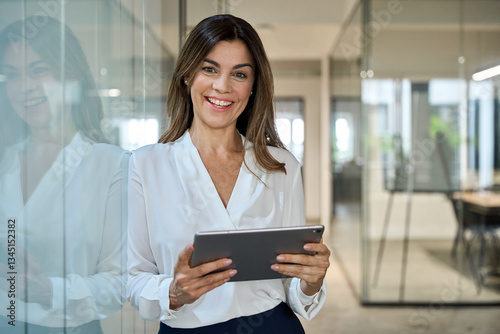Smiling mature business woman executive, happy middle aged businesswoman entrepreneur, 40 years old company hr holding digital tablet looking at camera standing in office at work. Portrait.