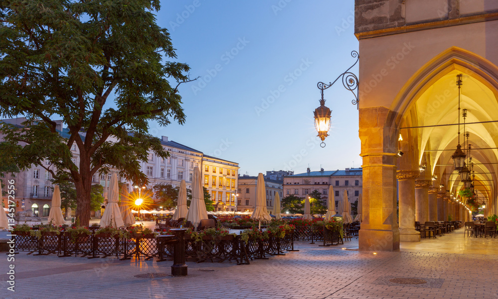 Evening view of the Main Square in Krakow, Poland, featuring the illuminated arcade of the Cloth Hall and outdoor cafes with closed umbrellas