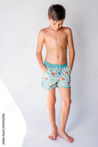 Handsome young beach boy wearing swim trunks standing with his hands in his pockets looking down at his feet