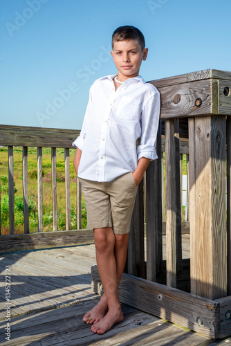 Smiling boy standing casually outside beach resort on a wooden deck, barefoot and wearing khaki shorts and a white poplin shirt