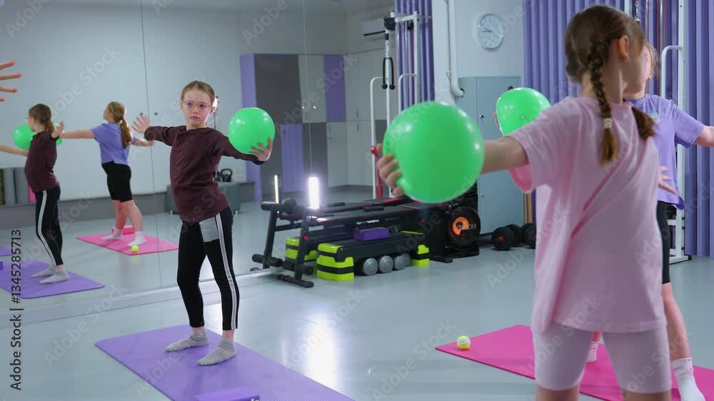 Children perform arm flexibility exercise with green balloons, gym equipment and music box visible in background of modern fitness center