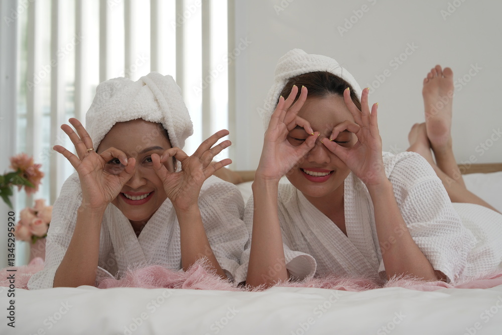 Asian mother and daughter in bathrobes laying on bed in bedroom, covering their eyes with their hands, having fun on a relaxing day, clean skin.