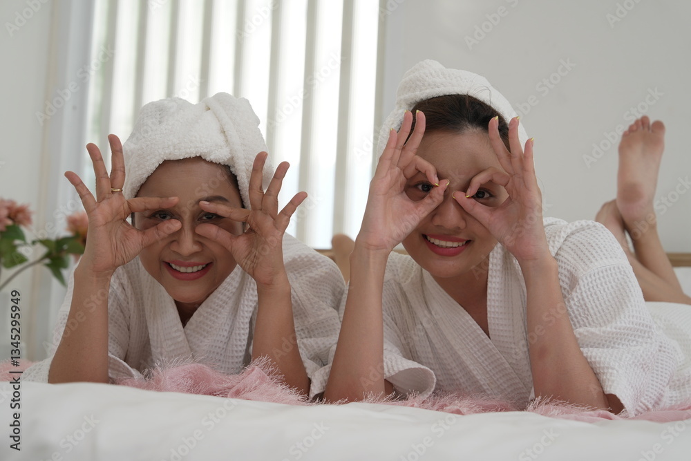 Asian mother and daughter in bathrobes laying on bed in bedroom, covering their eyes with their hands, having fun on a relaxing day, clean skin.