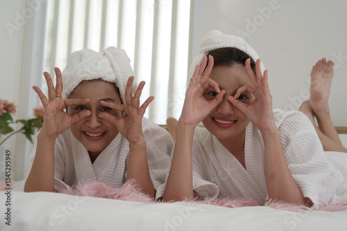 Obraz na płótnie Asian mother and daughter in bathrobes laying on bed in bedroom, covering their eyes with their hands, having fun on a relaxing day, clean skin. w salonie