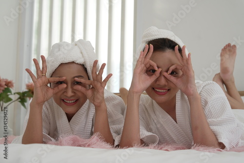 Obraz na płótnie Asian mother and daughter in bathrobes laying on bed in bedroom, covering their eyes with their hands, having fun on a relaxing day, clean skin. w salonie