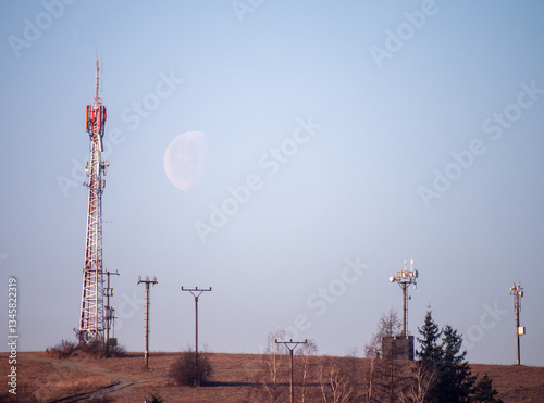 Rural landscape with telecommunications tower, power lines and visible moon in the sky. The scene depicts modern infrastructure blending with nature in the early morning.