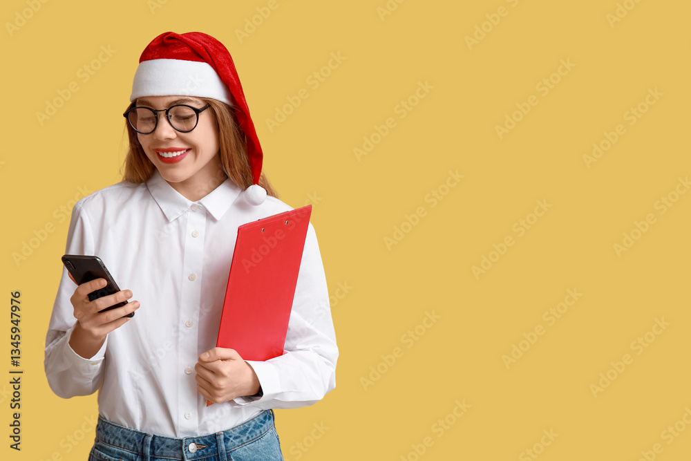 Young female real estate agent in Santa hat with mobile phone on yellow background Young female real estate agent in Santa hat with mobile phone on yellow background