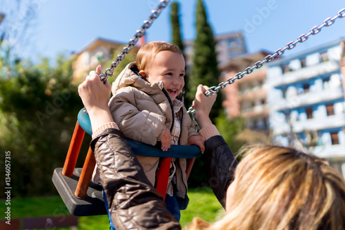 Mother pushing her happy baby boy on a swing in a playground