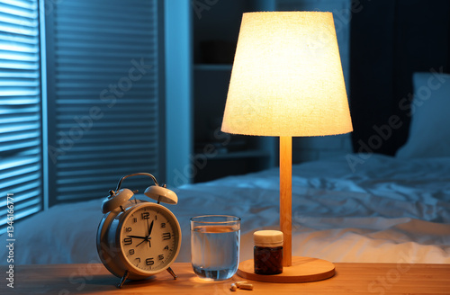 Alarm clock with melatonin pills, glass of water and glowing lamp on table in bedroom at night photo