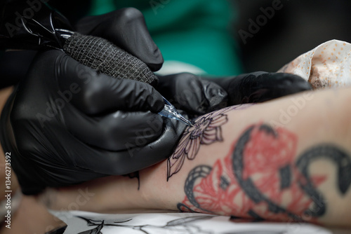 Close up tattoo master professional tattoo artist in black gloves making a rose tattoo on a man's forearm. He is holding a tattoo machine.
