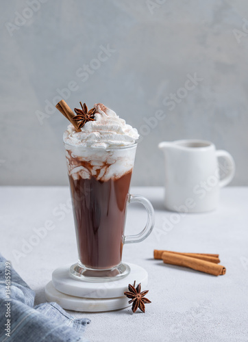 Hot chocolate with whipped cream, cinnamon and spices in a tall glass on a light background with ilk jug.