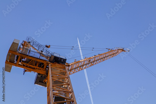 A towering construction crane set against a clear blue sky with a visible airplane contrail. The perspective emphasizes 