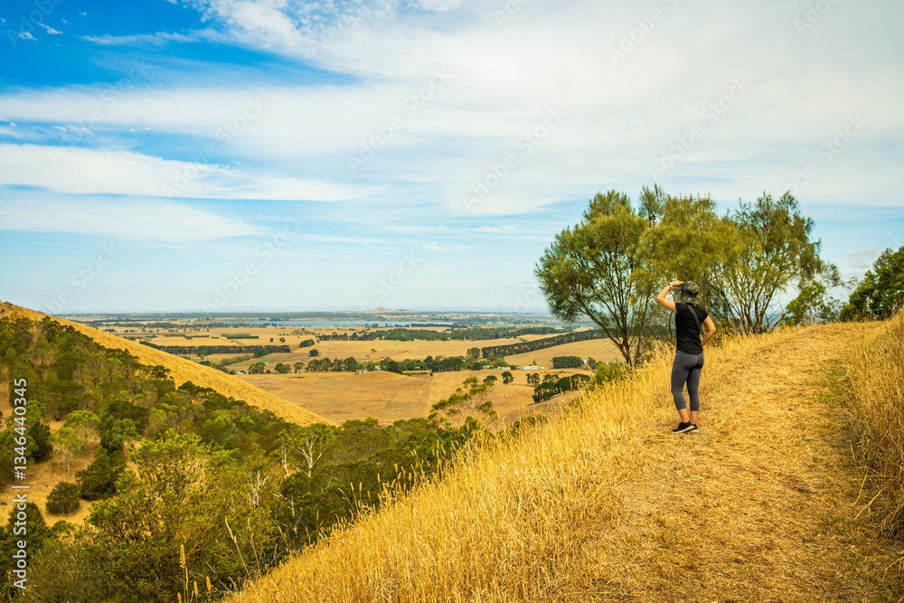 Fototapeta premium Rear view of woman standing on walking trail in a hill, overlooking the view in the far distance, Mount Sugarloaf, Camperdown, Victoria, Australia