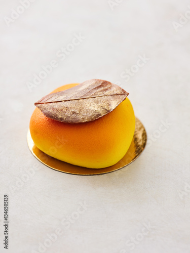 An elegant dessert shaped like a fruit sits on a gold plate, adorned with a metallic leaf. The vibrant colors of yellow and gold create a stunning presentation ideal for special occasions