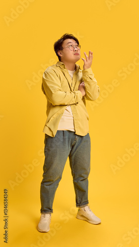 Full length photo of thoughtful, dreamy young Asian man, looking up with questionable expression against sunny yellow background. Concept of human emotions, idea, question, creative. Ad