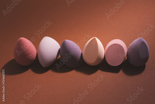 Egg-shaped makeup sponges, beauty blenders arranged in a row on a brown background. Dramatic lighting, deep shadows, stylish and artistic composition with beauty tools.