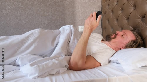 Young attractive man uses smartphone to watch videos and masturbates under the blanket in bed satisfying his sexual desire on his own. Porn addiction and excess hormone.