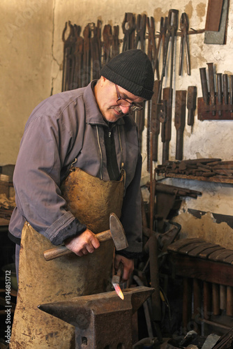 Blacksmith at work, mature man hitting a red-hot iron with a hammer on the anvil. Blacksmith forging red-hot metal bar in smithy