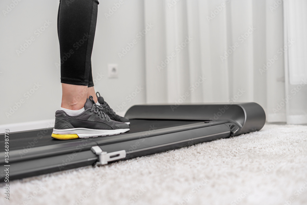 A person wearing black leggings and grey sneakers, with yellow soles is walking on a modern treadmill placed on a soft carpet in a minimalistic home setting.