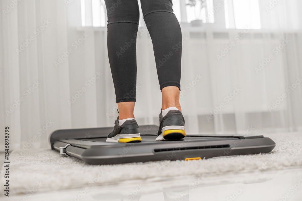 A person wearing black leggings and grey sneakers, with yellow soles is walking on a modern treadmill placed on a soft carpet in a minimalistic home setting.