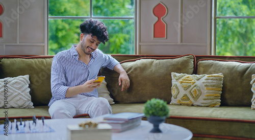 Canvas Print Indian happy young adult gen z man guy sitting cozy sofa couch hold mobile look
