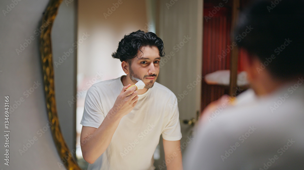 © Raushan_films - Indian Asian gen z young adult smiling guy bearded man look mirror hold soft brush apply white foam gel cream to his beard face before shave do morning day self skin care routine indoor house