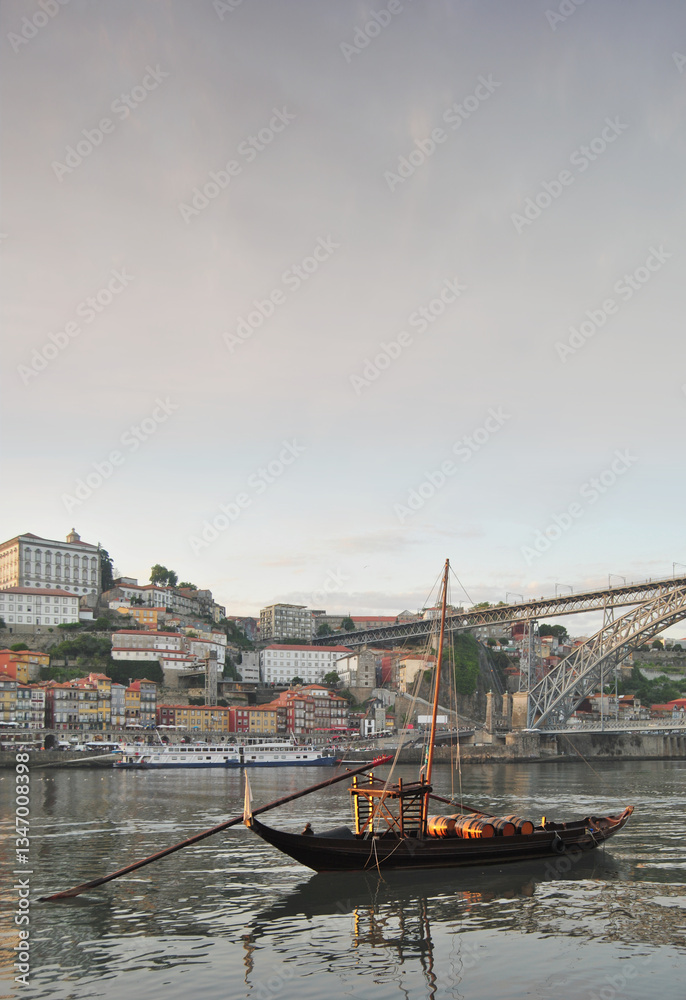 Obraz premium Portrait of downtown Porto in the late afternoon with a sunset sky, the Douro River with a traditional and old wine transport boat, part of the metal bridge of the Dom Luis metro bridge