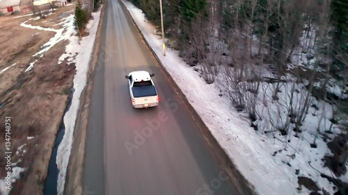 A truck travels down a gravel road surrounded by bare trees and patches of lingering snow. A small creek flows alongside, creating a serene winter atmosphere.