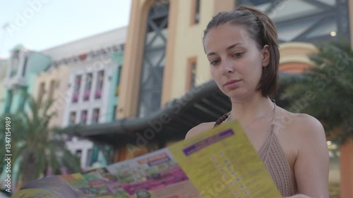A young woman in a summer outfit carefully reads a colorful park guide while standing in a vibrant amusement park filled with attractions. A young woman holds a park map
