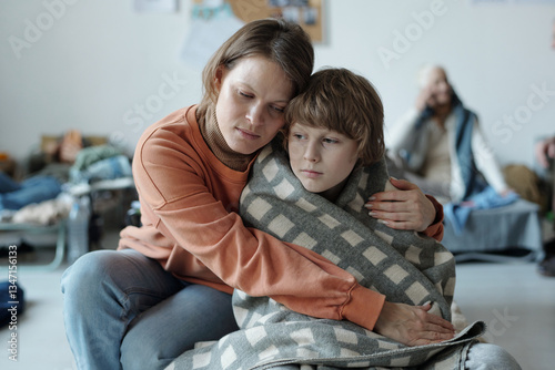 Careful young mother embracing her cute son wrapped in plaid while sitting close to him on sleeper in front of camera