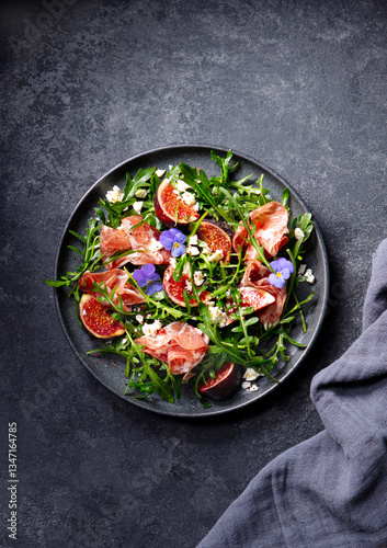 Arugula salad with fresh figs and prosciutto, jamon and edible flowers in a plate. Restaurant serving, plating. Dark background. Top view.