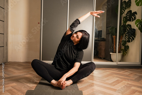 In the background is a wardrobe and a linoleum floor. A young woman in black sportswear is practicing yoga on a black mat in her room. The girl is in the lotus position.