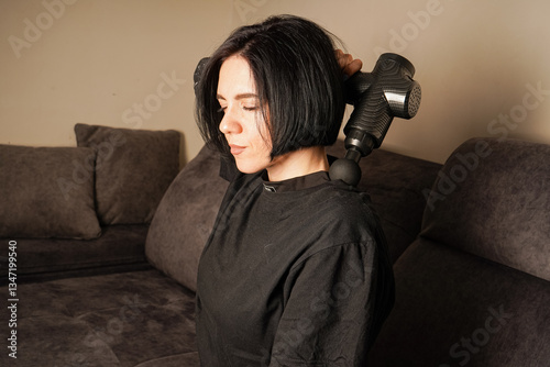 Using a percussion massager, the woman targets her leg muscles in a quiet living room. Her casual black outfit complements the serene environment. Perfect depiction of modern relaxation tools.