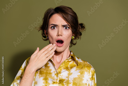 Surprised young woman with brunette hair in elegant blouse against khaki background, expressing astonishment with open mouth and wide eyes
