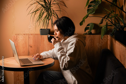 She holds a coffee cup and uses her laptop. The warm tones and casual attire create a calm and inviting scene. Sitting at a wooden table, the woman combines work and leisure.