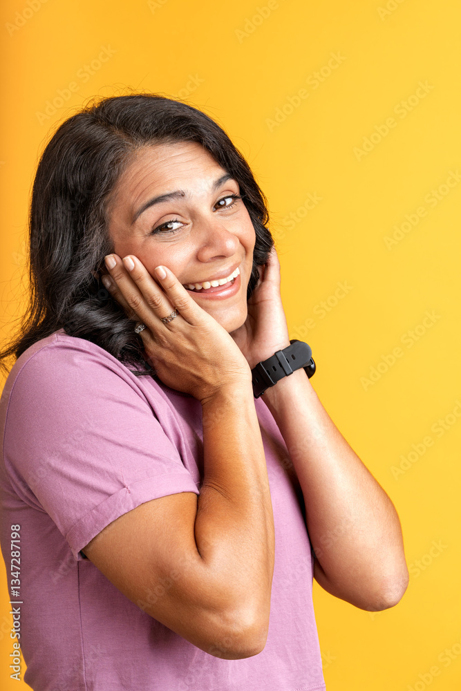 Fototapeta premium Excited latin woman expressing joy with hands on cheeks on yellow background