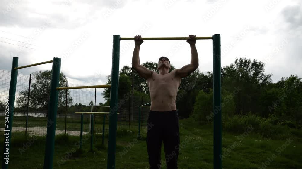 Muscular athlete doing pull ups on horizontal bar at sports ground. Young shirtless sportsman exercising at nature. Strong sporty man training outdoor. Concept of healthy and active lifestyle. Slow mo Muscular athlete doing pull ups on horizontal bar at sports ground. Young shirtless sportsman exercising at nature. Strong sporty man training outdoor. Concept of healthy and active lifestyle. Slow mo