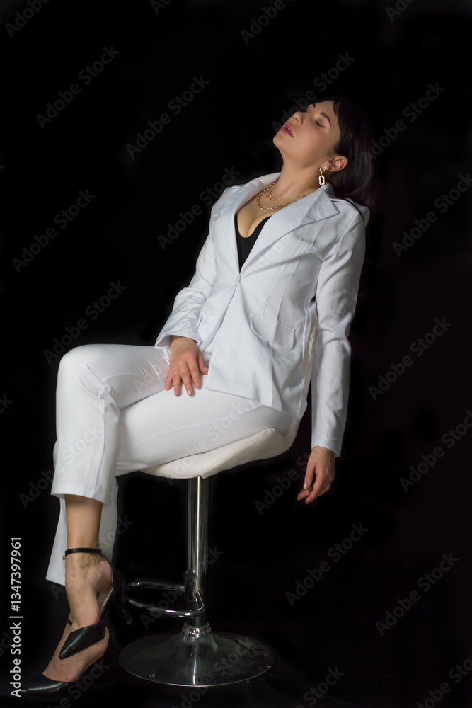Young tired woman in a black bra and a business white suit sits on a chair at a black background