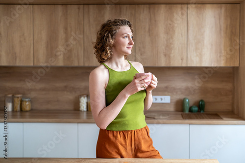 Serene woman stands in kitchen holds coffee mug takes mindful pause, reflecting on goals, affirmations. Relaxed young female in casual homewear enjoys quiet, smiles softly sets optimistic tone for day