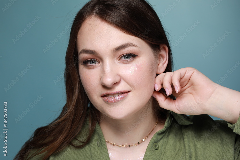 Naklejka premium A portrait of a young woman with glitter makeup. She smiles, her eyes expressive, and her lips covered in shimmer. Her hair flows in the wind, and the soft blue background adds freshness to the photo.