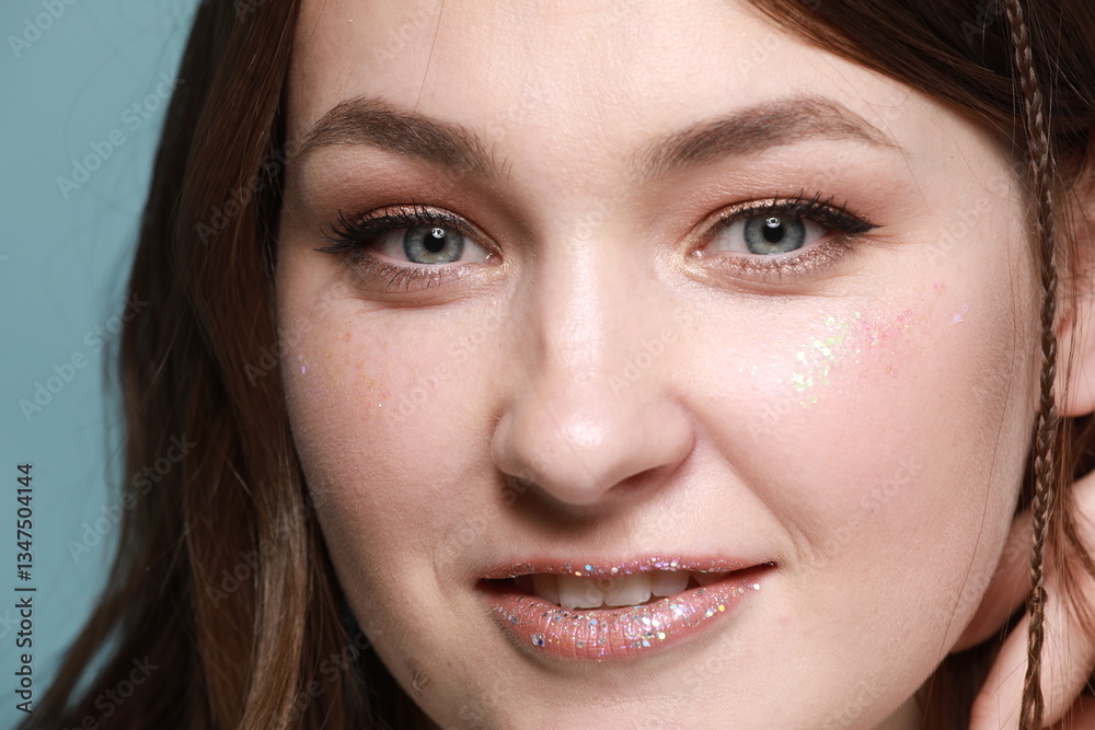 A portrait of a young woman with glitter makeup. She smiles, her eyes expressive, and her lips covered in shimmer. Her hair flows in the wind, and the soft blue background adds freshness to the photo. A portrait of a young woman with glitter makeup. She smiles, her eyes expressive, and her lips covered in shimmer. Her hair flows in the wind, and the soft blue background adds freshness to the photo.