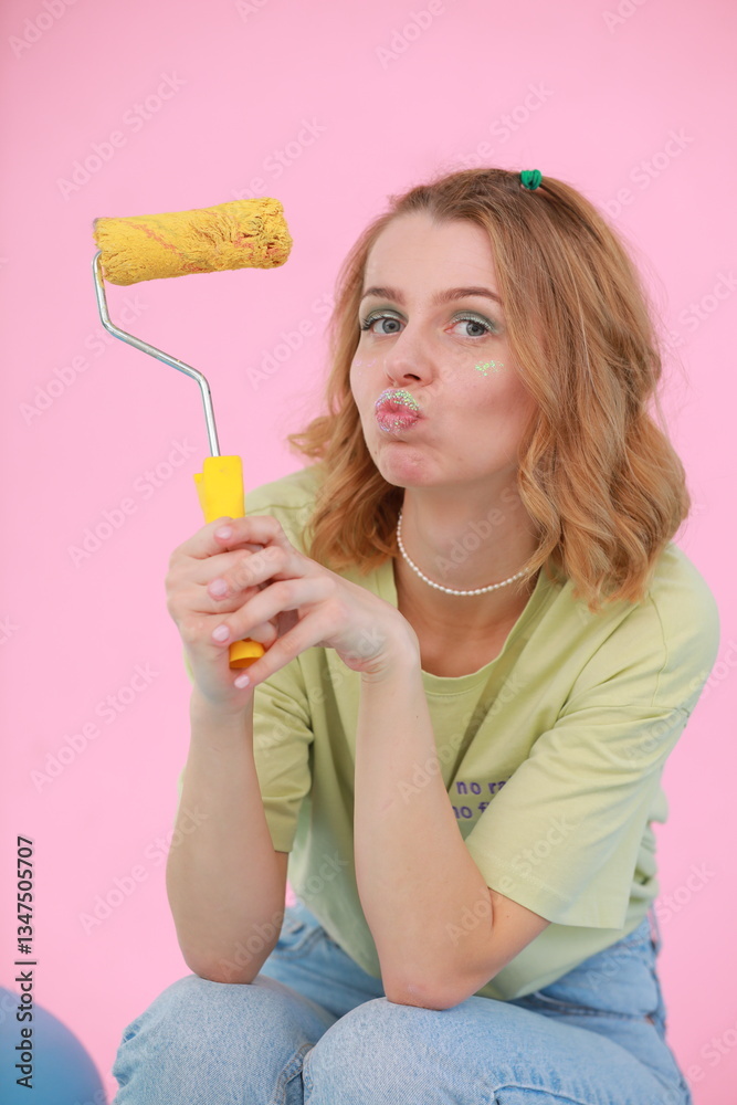 A young woman in a green T-shirt and jeans sits on a chair in a bright studio, playfully holding a large macaron-shaped pillow. Decor: pink background, big cactus, dessert-style props.