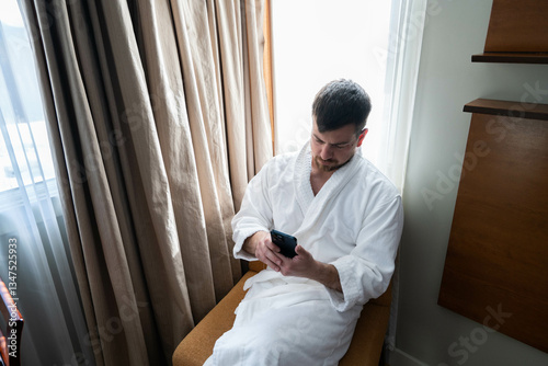 Man wearing white hotel bathrobe sitting on a chair near the window and checking incoming message on his smartphone