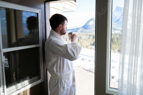 Young caucasian man wearing bath robe and drinking hot tea at his hotel room in the morning with fresh air and opened balcony Mountain View