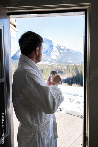 Young caucasian man wearing bath robe and drinking hot tea at his hotel room in the morning with fresh air and opened balcony Mountain View
