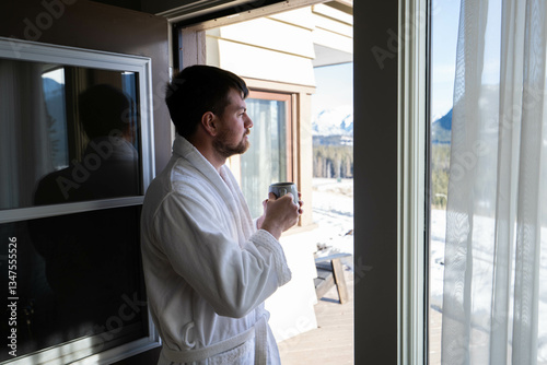 Young caucasian man wearing bath robe and drinking hot tea at his hotel room in the morning with fresh air and opened balcony Mountain View