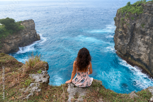 A beautiful woman in a pink dress sits on a cliff above the ocean on the island of Nusa Penida. Devil's Billabong an incredibly wonderful lagoon with splashes from the waves.